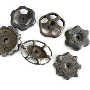 May include: A collection of six vintage metal valve handles in various shapes and designs. The handles are in shades of gray and brown, with visible wear and tear. Some handles have text that reads "OPEN" and "CLOSE".