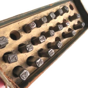 May include: A wooden box with a green lid containing a set of metal letter punches. The punches are arranged in rows and columns, and each punch has a different letter or symbol.