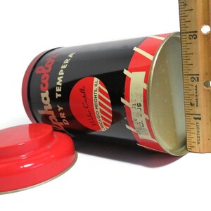 May include: A red and black tin can with a white label that reads "Alphacolor Dry Tempera" and "Waber Castella Chicago Heights, Ill." The can is open and the inside is white. A ruler is next to the can.