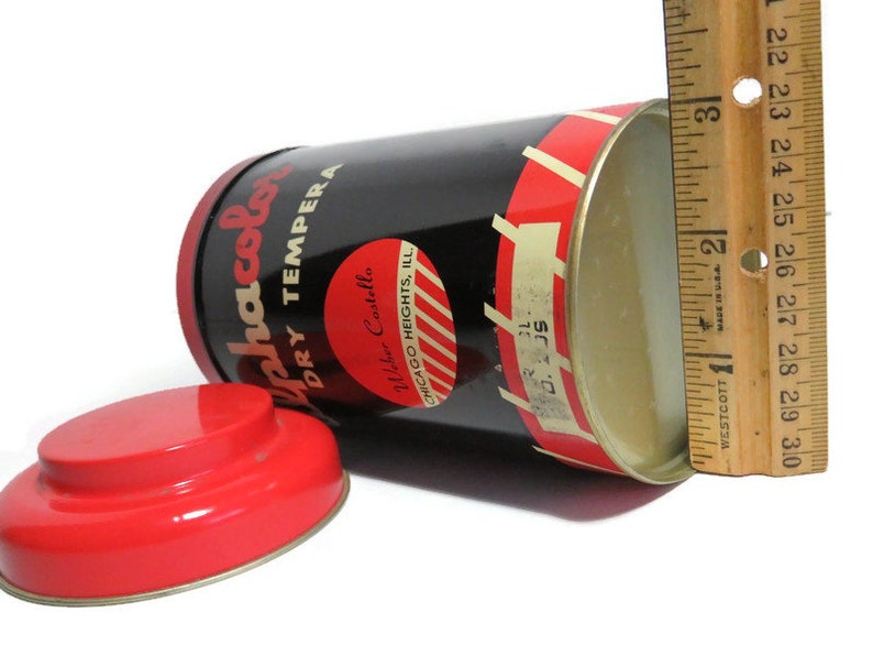May include: A red and black tin can with a white label that reads "Alphacolor Dry Tempera" and "Waber Castella Chicago Heights, Ill." The can is open and the inside is white. A ruler is next to the can.