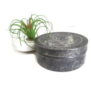 May include: A round, antique metal tin with a weathered, gray finish. The lid features embossed lettering. A small, white pot with a green plant is next to the tin.