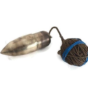 May include: A metallic, bullet-shaped plumb bob with a pointed tip and a darkened finish, connected by a thin cord to a spherical ball of brown string that is secured with a bright blue rubber band against a plain white background.