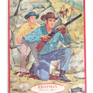 May include: Vintage jigsaw puzzle featuring a scene from the TV show "The Rifleman". The puzzle depicts Chuck Connors as Lucas McCain, kneeling and aiming a rifle, with his son Mark McCain standing behind him. The puzzle box is red with the title "Rifleman" and the words "Frank  May Puzzles" printed on it.