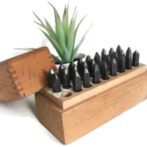 May include: A wooden box with a set of 30 black metal letter stamps. The box is open and the stamps are arranged in rows. The box is made of wood and has a light brown color. The stamps are made of metal and have a black color.