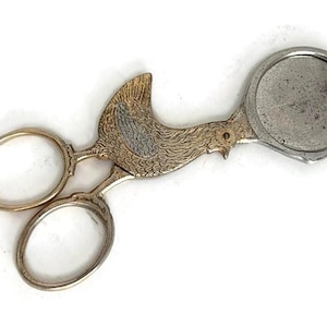 May include: A metal egg topper tool featuring a gold-toned handle shaped like a bird with textured feather details. The tool has two oval finger loops and a circular silver-toned cutting cup at the end designed to remove the top of an egg shell.