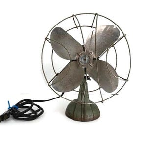 May include: An antique, four-blade electric fan with a metal cage and base. The fan blades and cage have a weathered, silver finish. The base is a muted green color, and the fan has a black power cord.