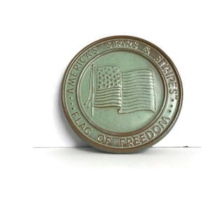 May include: A round, antique-style coin with a green and bronze finish. The coin features an embossed American flag and the text "America's Stars & Stripes Flag of Freedom" around the edge. The coin is set against a white background.