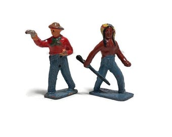 May include: Two vintage metal toy figurines. One is a cowboy in a red shirt and blue jeans, holding a gun. The other is a Native American figure in a red shirt and blue jeans, holding a weapon. Both figures are on blue bases.