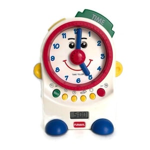 May include: A white and colorful PlaySkool Time Teller clock with a red and white clock face, blue hands, and a digital display showing 5:00. The clock has a green "TIME" label and blue feet.