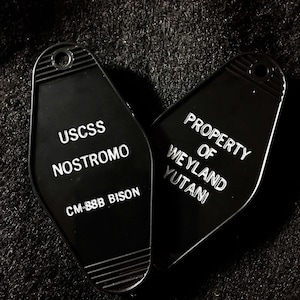 May include: Two black keychains with white text. The keychain on the left says "USCSS Nostromo CM-88B Bison". The keychain on the right says "Property of Weyland Yutani".