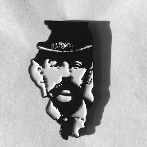 May include: Black and white enamel pin shaped like the state of Illinois with a silhouette of a man wearing a hat and a beard.