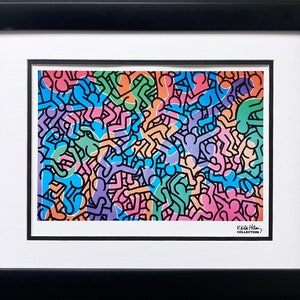 Op de afbeelding: Een ingelijste print met een levendig kunstwerk met een herhalend patroon van gestileerde figuren in verschillende kleuren, waaronder blauw, groen, oranje en roze. Het kunstwerk is ingelijst in een zwart frame met een witte passe-partout. De print is gelabeld "Keith Haring Collection."