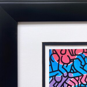 Op de afbeelding: Ingelijst kunstwerk met een kleurrijk abstract ontwerp met figuren in blauw, roze, groen en paars, omlijnd in zwart. Het kunstwerk is gevat in een zwarte lijst met een witte passe-partout, wat een moderne uitstraling cre&euml;ert.