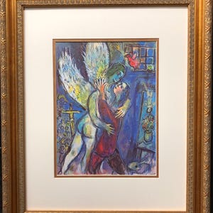 Puede incluir: Una litografía a color de una pintura de Marc Chagall que representa a un ángel alado abrazando a un hombre con una túnica roja. El fondo es de un azul vibrante con una ventana y un jarrón de flores.