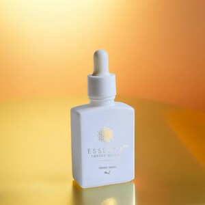 May include: A white dropper bottle with a square base and a rounded top. The bottle has gold text that reads "ESSENCE 54 EMBODY DIVINE" and "SIDERIC DROPS". The background is a gradient of gold and orange.
