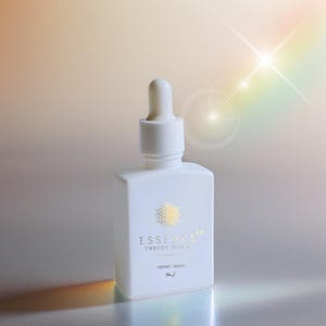 May include: A white, rectangular bottle with a dropper top. The bottle has gold text that reads "ESSENCE EMBODY DIVINE" and "SIDERIC DROPS". The bottle is on a reflective surface with a soft, blurred background.