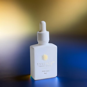 May include: A white, square-shaped bottle with a dropper top. The bottle has gold text that reads "ESSENCE64 EMBODY DIVINE" and "SIDERIC DROPS". The background is a gradient of gold and blue.