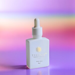 May include: A white, square-shaped bottle with a dropper top. The bottle has gold text and a gold design. The text on the bottle reads "ESSENCE EMBODY DIVINE" and "SIDERIC DROPS". The background is a gradient of pink, purple, and orange.
