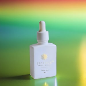 May include: A white, square-shaped bottle with a dropper top. The bottle has gold text and a gold emblem. The text reads "ESSENCE E64 EMBODY DIVINE SIDERIC DROPS 50ml". The background is a gradient of green, yellow, and red.