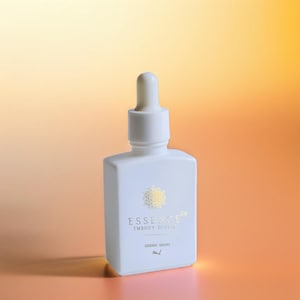 May include: A white dropper bottle with a square base and a rounded top. The bottle has gold text that reads "ESSENCE", "EMBODY DIVINE", and "SIDERIC DROPS". The background is a gradient of orange and yellow.