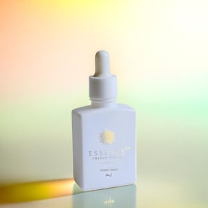 May include: A white, square-shaped bottle with a dropper top. The bottle has gold text that reads "ESSENCE 64 EMBODY DIVINE" and "SIDERIC DROPS". The background is a gradient of yellow, green, and pink.