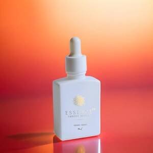 May include: A white, square-shaped bottle with a dropper top. The bottle has gold text that reads "ESSENCE EMBODY DIVINE SIDERIC DROPS 5mL". The background is a gradient of red and orange.