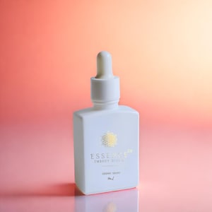 May include: A white, square-shaped bottle with a dropper top. The bottle has gold text that reads "ESSENCE EMBODY DIVINE" and "SIDERIC DROPS". The background is a gradient of pink and orange.