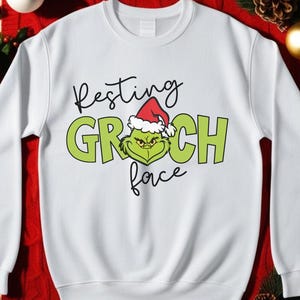 May include: White sweatshirt with a green Grinch face wearing a Santa hat and the text "Resting Grinch Face".