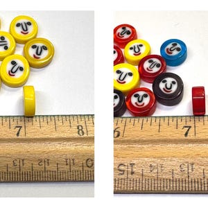 May include: A collection of colorful, round beads with smiling faces. The beads are yellow, red, blue, and brown. A ruler shows the beads are approximately 0.25 inches thick. These beads are suitable for jewelry making or craft projects.