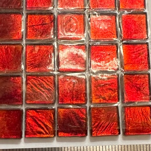 May include: A sheet of 25 small, square, red iridescent glass tiles. The tiles are approximately 1 inch by 1 inch. A ruler is in the bottom of the image to show the size of the tiles.
