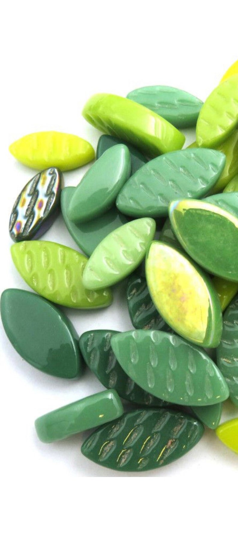 May include: A collection of green leaf-shaped glass beads in various shades, including light and dark greens, and a few with textured surfaces. Some beads have a yellow hue.