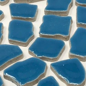 May include: A collection of irregularly shaped, glossy blue ceramic tiles with grey edges. The tiles are arranged on a white surface. The tiles have a smooth, reflective surface and a slightly uneven shape, suitable for mosaic or decorative use.