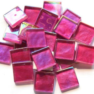 May include: A collection of square, iridescent glass tiles in shades of pink and purple. The tiles have a glossy finish and are arranged in a scattered pile. The edges of the tiles reflect a rainbow of colors.