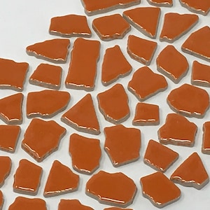May include: A collection of orange ceramic mosaic tiles in various irregular shapes. The tiles are arranged in a random pattern on a white background.