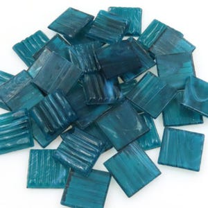 May include: A collection of square, teal-colored glass tiles. The tiles have a textured surface with varying patterns, creating a mosaic effect. The tiles are scattered, with some showing a ribbed design and others a smooth finish.