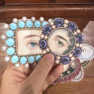 May include: A hand holding several decorative frames with images of eyes. One frame is rectangular with turquoise stones and clear gems. Another is round with purple gems and pearls. The frames are on a wooden surface.