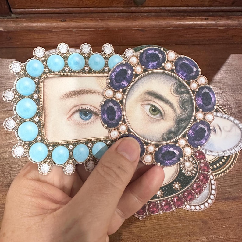 May include: A hand holding several decorative frames with images of eyes. One frame is rectangular with turquoise stones and clear gems. Another is round with purple gems and pearls. The frames are on a wooden surface.