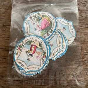 May include: A stack of vintage, round, blue and white paper labels with illustrations and text. The labels feature a woman in a pink dress, a woman in a red dress, and a woman in a blue dress. The text on the labels includes "Extrait de F.d'Italic", "Extrait de Muse", "DE DISSEY et PIVER A PARIS", "Rue Martin", and "H&eacute;liotrope".