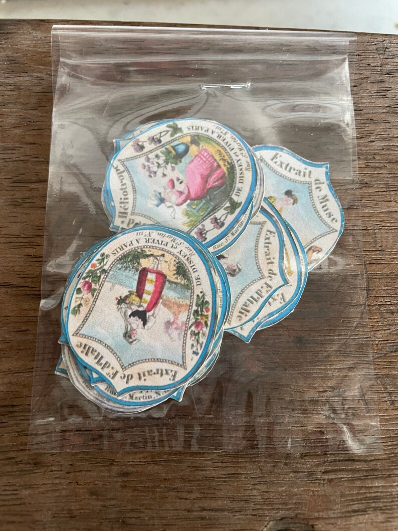 May include: A stack of vintage, round, blue and white paper labels with illustrations and text. The labels feature a woman in a pink dress, a woman in a red dress, and a woman in a blue dress. The text on the labels includes "Extrait de F.d'Italic", "Extrait de Muse", "DE DISSEY et PIVER A PARIS", "Rue Martin", and "H&eacute;liotrope".