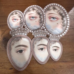 May include: A collection of antique-style oval and heart-shaped portraits. The portraits feature detailed illustrations of eyes and faces, some with crowns. The color palette includes soft pinks, blues, and browns, with a pearl-like border.