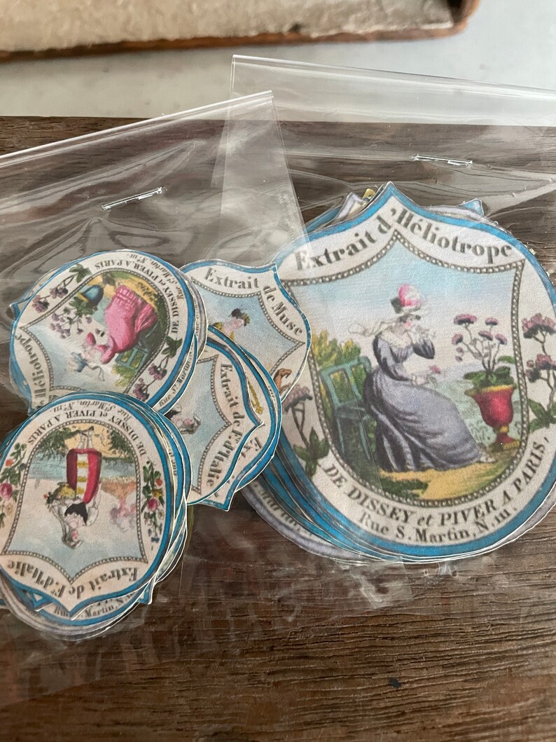 May include: A stack of vintage paper labels with blue and white borders. The labels feature illustrations of women in dresses and the text "Extrait de Heliotrope" and "DE DISSEY et PIVER A PARIS, Rue S. Martin, N.11."