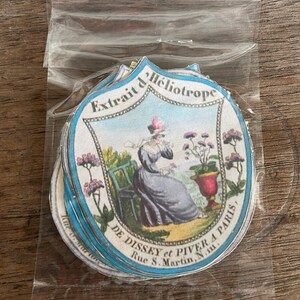 May include: A stack of vintage round labels with a blue and white border. The label features a woman in a blue dress sitting in a chair with a potted plant. The text on the label reads "Extrait d'Heliotrope DE DISSEY et PIVER A PARIS, Rue S. Martin, N.m."