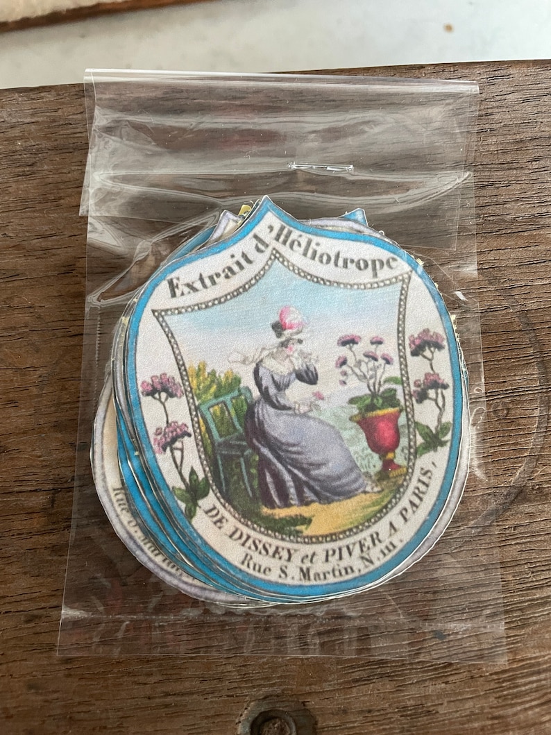 May include: A stack of vintage round labels with a blue and white border. The label features a woman in a blue dress sitting in a chair with a potted plant. The text on the label reads "Extrait d'Heliotrope DE DISSEY et PIVER A PARIS, Rue S. Martin, N.m."