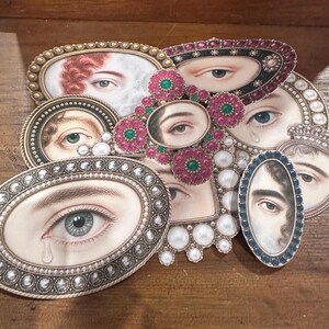 May include: A collection of oval and rectangular brooches and pendants featuring painted eye portraits. The frames are embellished with pearls, rhinestones, and colored gemstones. The color palette includes silver, gold, pink, green, and blue.