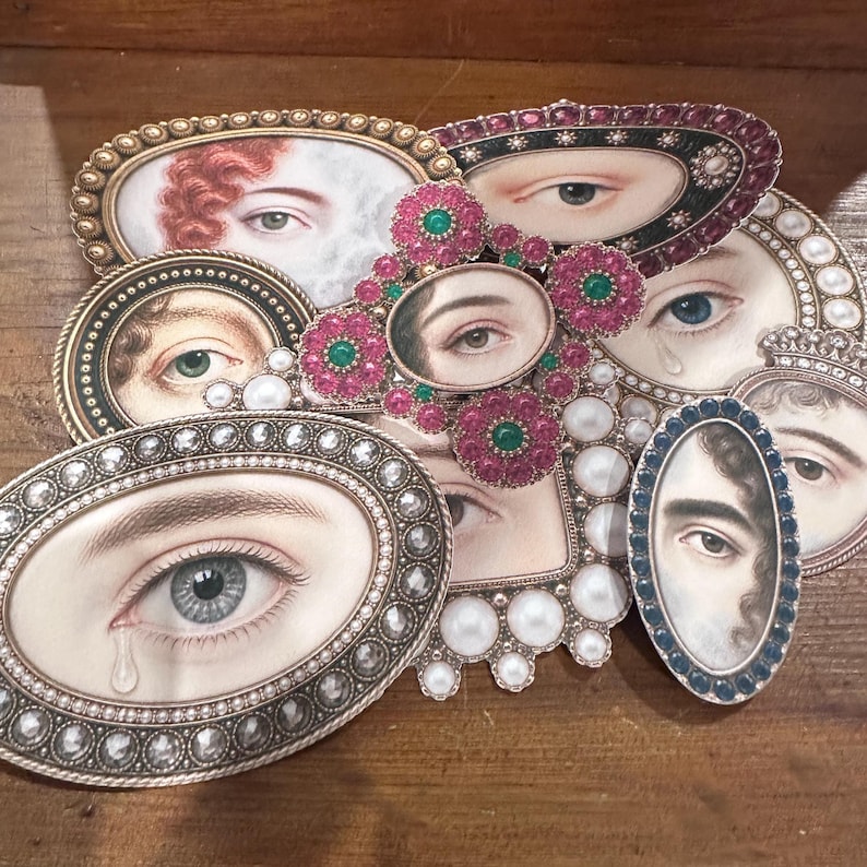 May include: A collection of oval and rectangular brooches and pendants featuring painted eye portraits. The frames are embellished with pearls, rhinestones, and colored gemstones. The color palette includes silver, gold, pink, green, and blue.
