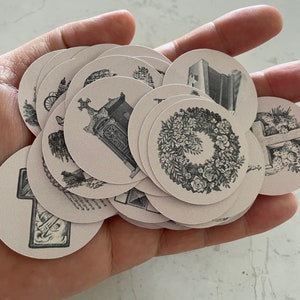 May include: A collection of round, light pink stickers with black and white vintage illustrations. The stickers feature images of a wreath, a cross, a carriage, and architectural elements. The stickers are held in a hand.