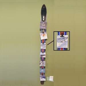 May include: A repurposed ski display featuring a black and white ski with multiple clips holding various cards and brochures. The cards have images and text, including "Snowbasin Winter Trail Map & Mountain Guide". The display is mounted on a light green wall.