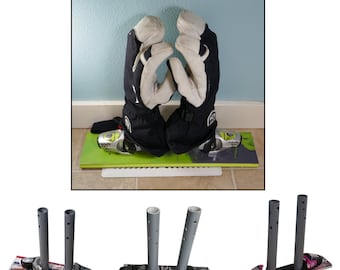 May include: A pair of black and white ski gloves resting on a green and gray snowboard. The gloves are upright, with the palms facing each other. The snowboard is on a white base, and the background is a light blue wall.