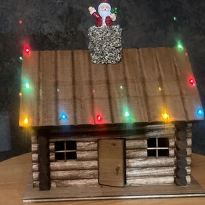 May include: A wooden log cabin model featuring a textured stone chimney with a small Santa Claus figurine on top. The roof is decorated with multi-colored LED string lights in red, green, blue, and yellow. The cabin has a central door and two windows.