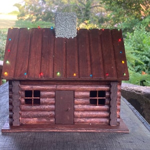 May include: A wooden log cabin model with a brown roof and a chimney. The roof is decorated with colorful lights.